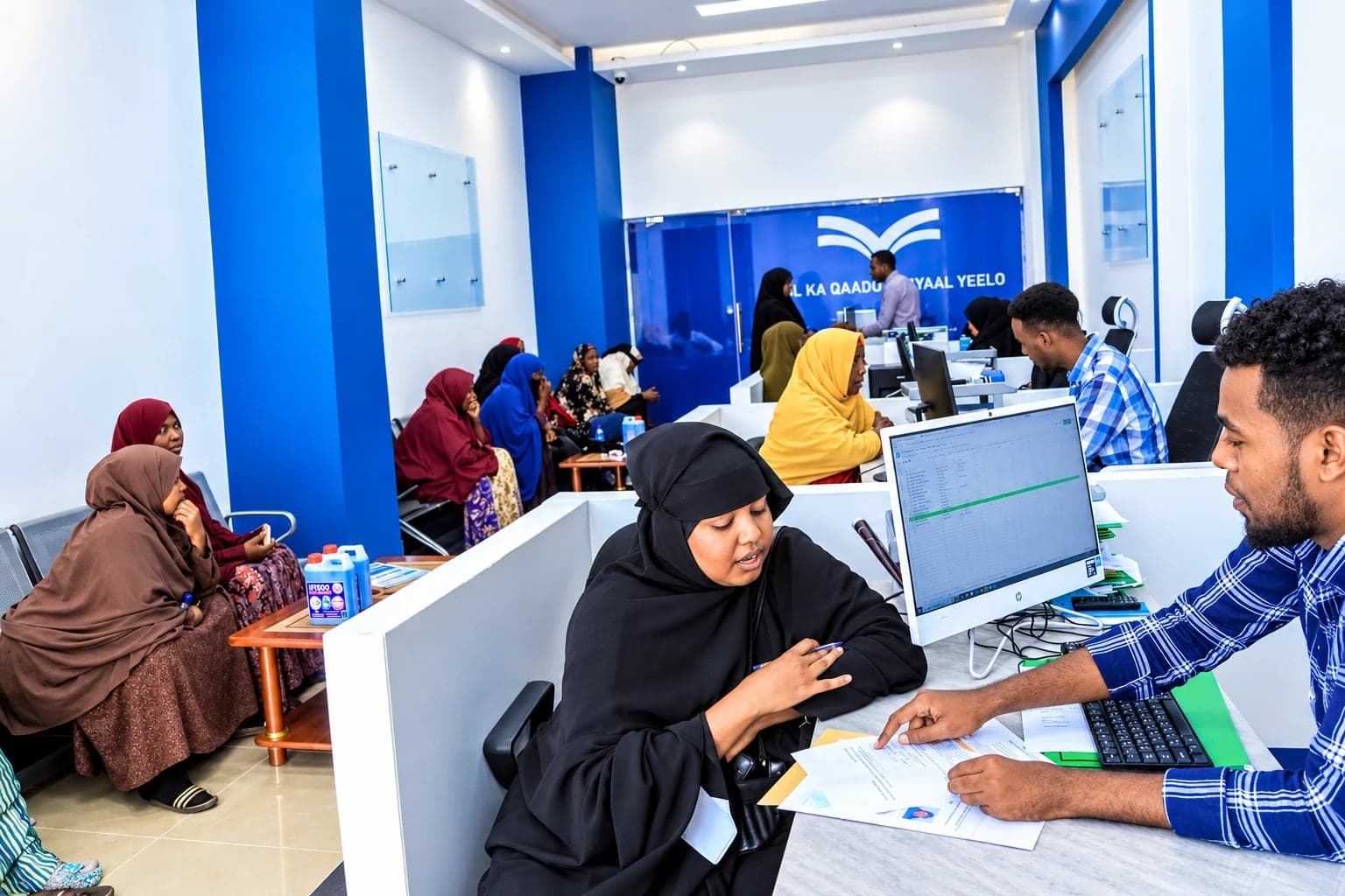 A Maal Microfinance officer helps a customer at a branch service desk while others wait